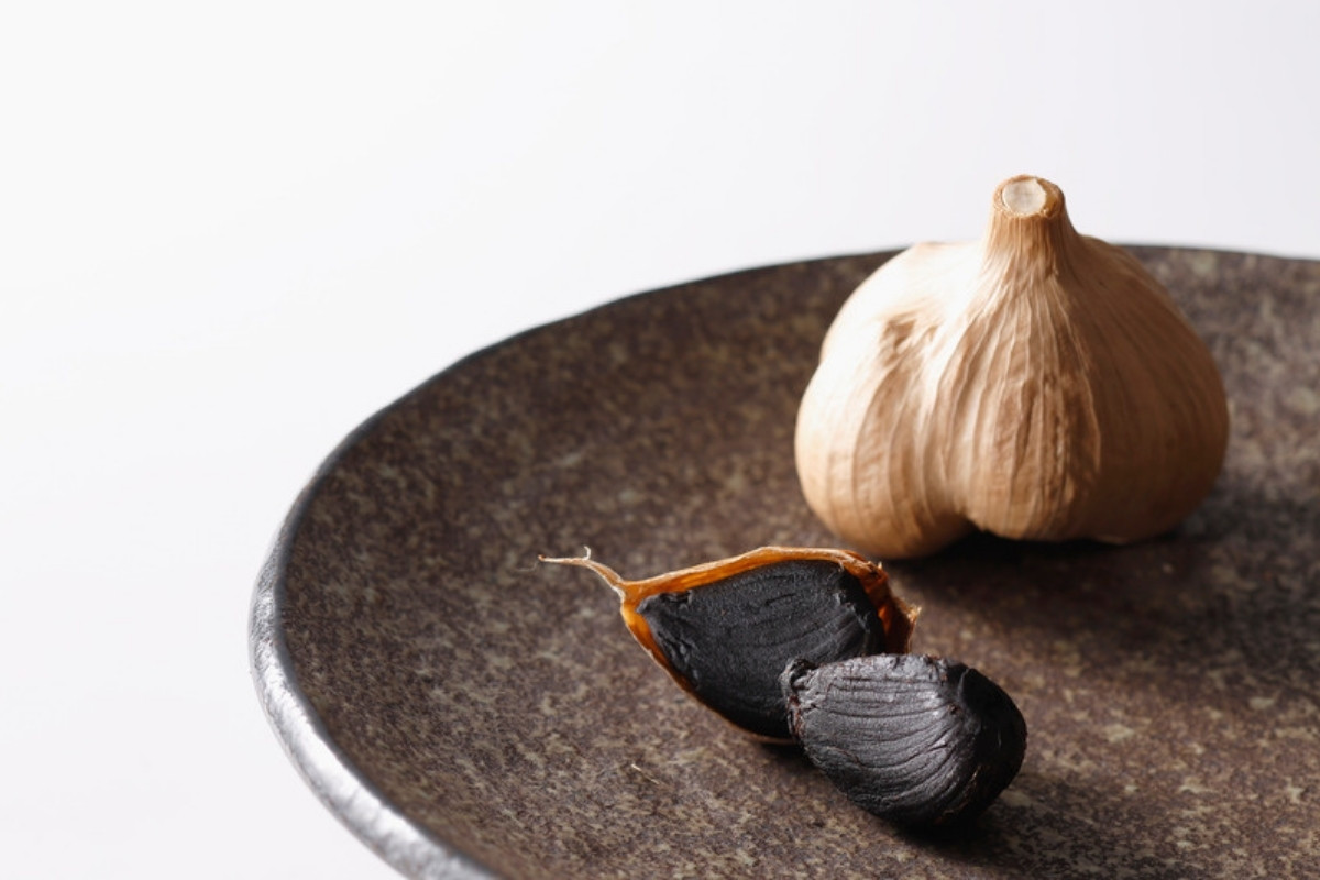 Black garlic cloves beside a whole white garlic bulb on a dark stone plate