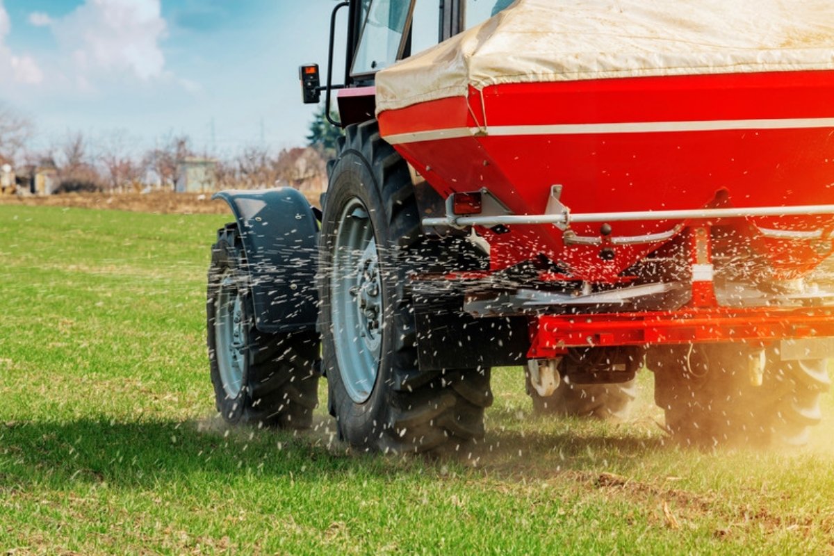 Épandeur rouge porté par un tracteur projetant des granulés d'engrais sur un champ vert