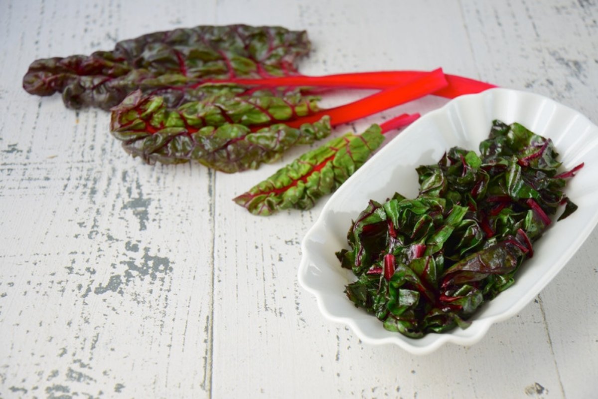 Fresh red-stemmed Swiss chard leaves next to a white bowl of cooked chard