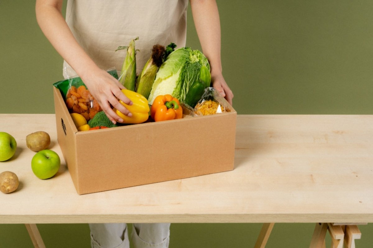 Hands packing fresh vegetables and fruit into a cardboard box for delivery